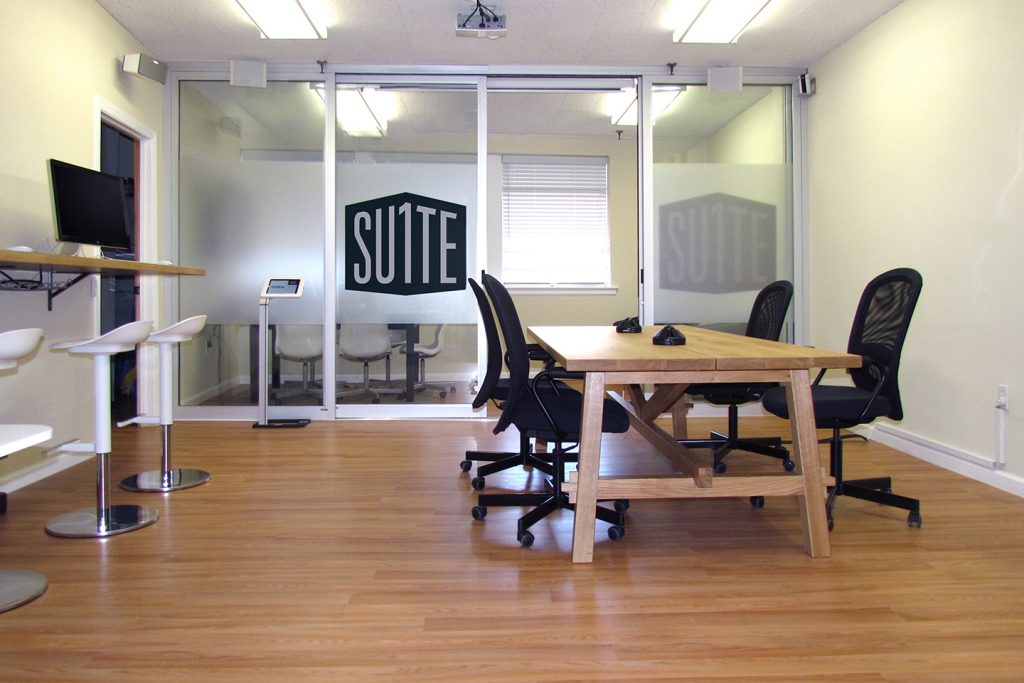 Suite1 Office in Millburn, NJ | An indoor view of an office with a long, wall-mounted desk and bar stools, a lower large table with office chairs, and a glass-walled conference room with a large Suite 1 Logo on the door.