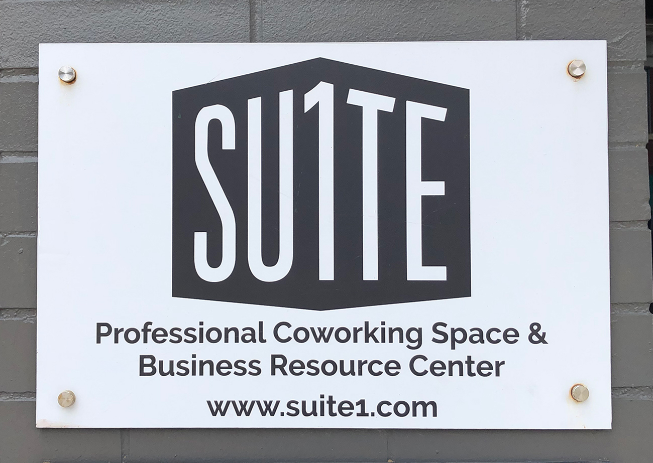A white sign mounted with silver pegs against a gray-painted brick wall. On the sign is Suite 1 Logo | A black polygon on white ground with white letters that say SU1TE in a sans-serif font.