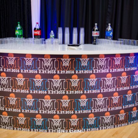 Curved bar with bar mitzvah logo in repeat pattern on front, sodas and plastic cups on bar top.